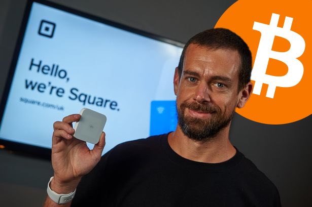 A man with short hair and a beard wearing a black shirt holds a small square gray device in his right hand while standing in front of a large screen displaying the Square logo, the text Hello were Square square.com, and a Bitcoin symbol overlay in orange circle.