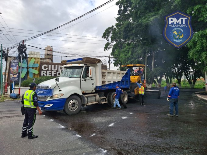 A white dump truck with blue accents is parked on a wet road surface during asphalt work, with its bed raised and gravel spilling onto the ground. Yellow construction machinery is nearby, and several workers in blue uniforms and safety gear stand around directing the operation. Police officers in green vests monitor the scene. In the background, a large sign reads CIUDAD REFORMADORES with floral decorations and a PMT VILLA NUEVA badge, surrounded by green trees, power lines, and a cloudy sky.