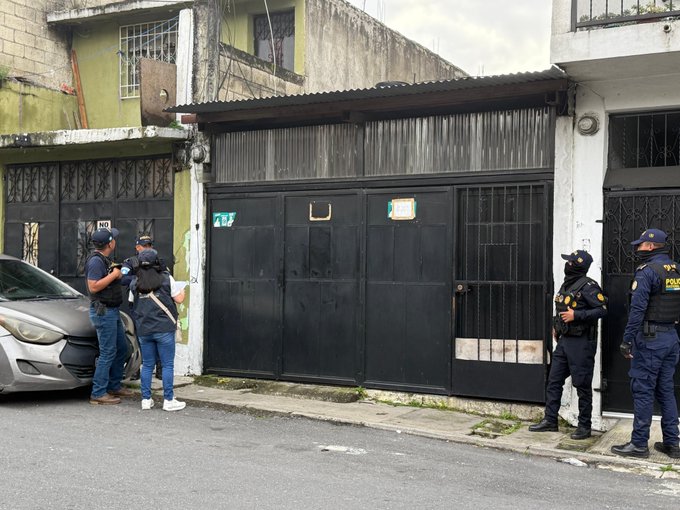 Several police officers in blue uniforms and tactical gear stand outside a black metal gate of a building in an urban neighborhood with green and gray walls, barred windows, and graffiti. A silver Hyundai car is parked nearby with doors open. Officers carry backpacks and equipment, positioned in a group discussing or preparing for an operation. The scene includes a dirt road, streetlight, and overcast sky.