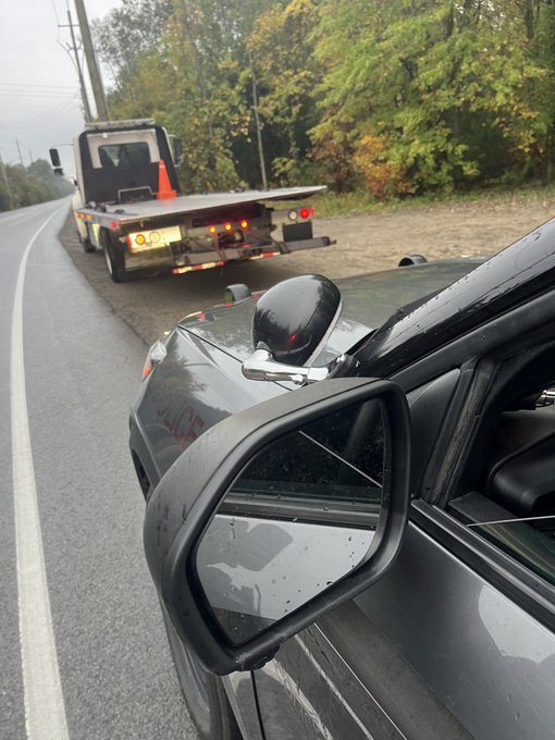 Roadside scene on a wet paved road with autumn foliage and utility poles in the background under overcast skies. A yellow tow truck with orange traffic cone and flashing lights tows a black sedan with a glossy black side mirror visible. The tow truck has a closed cargo area and is positioned behind the sedan on the shoulder.