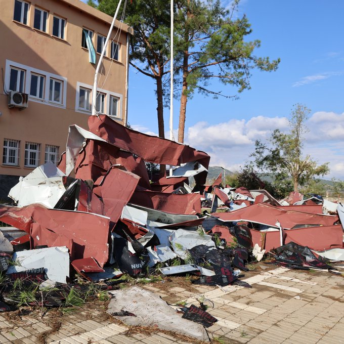 First image shows a beige school building with a partially torn-off roof scattered with metal sheets and debris on the ground surrounded by pine trees and a Turkish flag in a rural mountainous area. Second image depicts piles of red and gray metal roofing fragments and debris scattered across a paved courtyard next to the same school building with trees and a flagpole under a partly cloudy sky.