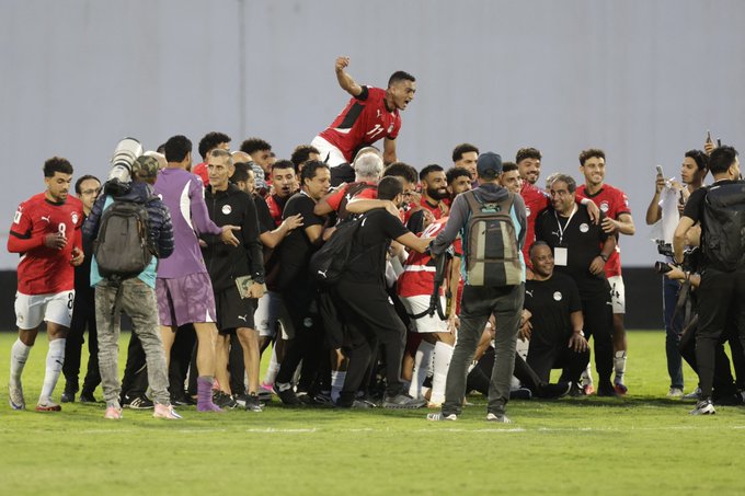 Un grupo de futbolistas egipcios con camisetas rojas y pantalones cortos negros celebran en un campo de fútbol verde después de una victoria. Un jugador salta alto en el centro con los brazos en alto, rodeado de compañeros de equipo que lo abrazan y lo vitorean. Funcionarios y personal con diversos atuendos se paran cerca, algunos tomando fotos. La escena se desarrolla con un telón de fondo de estadio con una pared gris.