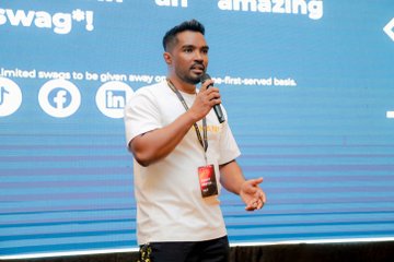 First image shows a man with short dark hair wearing a white t-shirt lanyard and red badge holding a microphone speaking on stage with blue backdrop text reading amazing and limited swags to be given away on first served basis social media icons below. Second image depicts a large diverse group of men and women in casual attire standing together smiling in front of a banner reading Binance Kandy Meetup with yellow Binance letters on floor and arched ceiling with warm lighting. Third image features five young men in casual clothes posing with cardboard signs reading Then Binance Now hands on yellow backdrop holding diamond hand and HODL always to the moon signs. Fourth image shows multiple men and women seated at a round table with glasses of orange drinks napkins and plates in a conference setting with warm lighting.