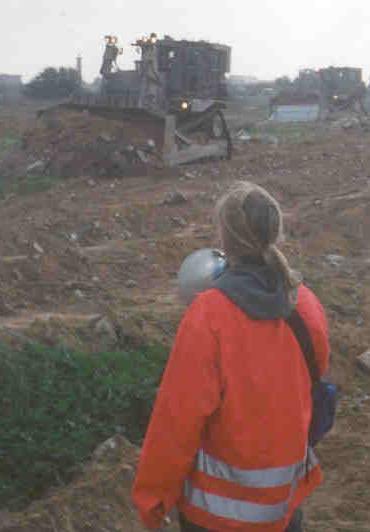 First image displays a bulldozer operating on a dirt mound in a barren area with industrial structures and another vehicle in the foggy background, viewed from behind a person wearing a red high-visibility jacket with gray hood, safety vest, ponytail, and carrying a gray bag. Second image shows a blonde woman with shoulder-length hair, smiling at the camera, wearing a white shirt, necklace with pendant, and pearl earrings, with another person partially visible on the side wearing green clothing and a necklace.