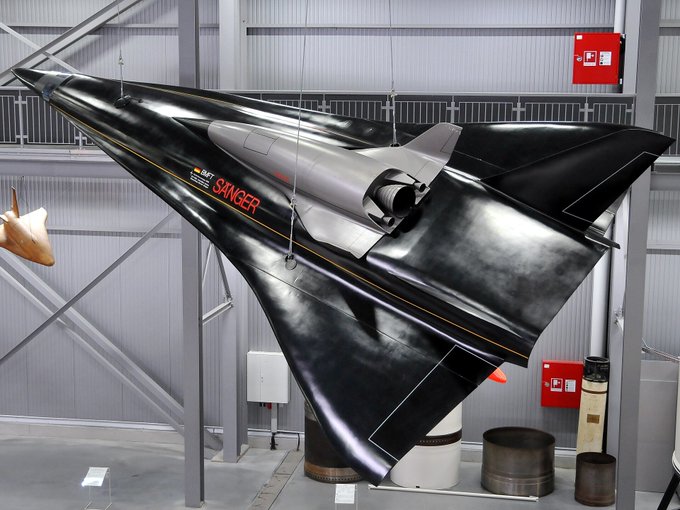 A detailed scale model of the Sänger II spaceplane, featuring a sharp delta-wing configuration in glossy black with red and white markings on the fuselage and wings, suspended from the ceiling in an industrial exhibit space with metallic beams, fire extinguishers on walls, and nearby cylindrical objects resembling rocket components or stands.