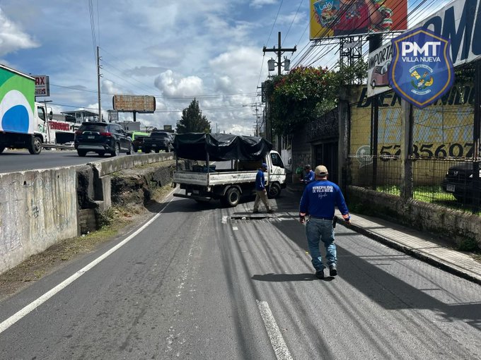 Two workers in blue jackets stand near a white truck covered with a blue tarp on a roadside, one holding a tool and the other walking away, surrounded by passing vehicles including green and white trucks, a bridge, utility poles, flowering plants, billboards with advertisements, and a sign displaying PMT VILLANUEVA logo under a partly cloudy sky.