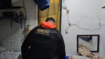 First image shows a police officer in black vest labeled POLICIA NACIONAL CIVIL standing in a room with shelves clothes and household items. Second image depicts an officer in black hooded jacket labeled DIPANDA INVESTIGACION CRIMINAL at a doorway with hanging clothes and blue walls. Third image features an officer in black clothing and vest labeled INTERPOL standing near a door with baskets and plastic items in a room with blue walls. Fourth image displays stacks of Guatemalan quetzale banknotes on the floor marked with evidence tag 1 next to a police officer in blue uniform wearing gloves pointing to the cash.