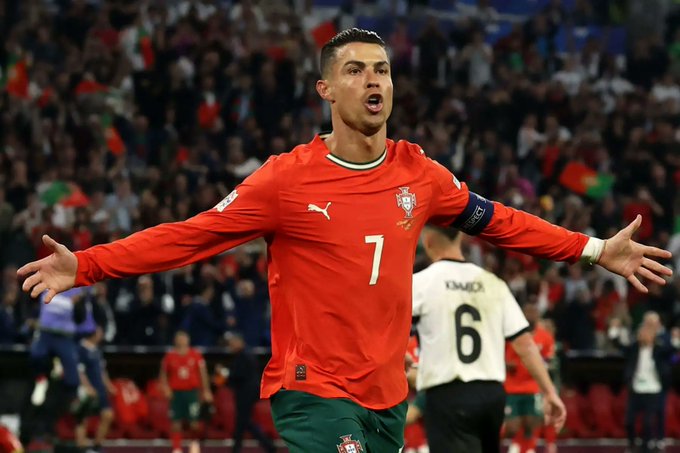 Cristiano Ronaldo se para en un campo de fútbol con una camiseta roja de manga larga de Portugal con el número 7 y el logotipo de PUMA, los brazos extendidos en celebración, la boca abierta como si gritara. Tiene el pelo corto y oscuro y una expresión concentrada. Detrás de él, se ve a un jugador con una camiseta blanca y negra de Alemania con el número 6. El estadio está lleno de espectadores en las gradas. El fondo muestra hierba verde y multitud borrosa.