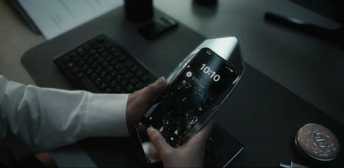 A hand in a white shirt sleeve holds a shattered silver iPhone displaying a lock screen with time 10:10 against a dark background on a wooden desk with papers, a keyboard, a black gadget, a metallic wedge-shaped object, a round black item with a logo, and a mouse.