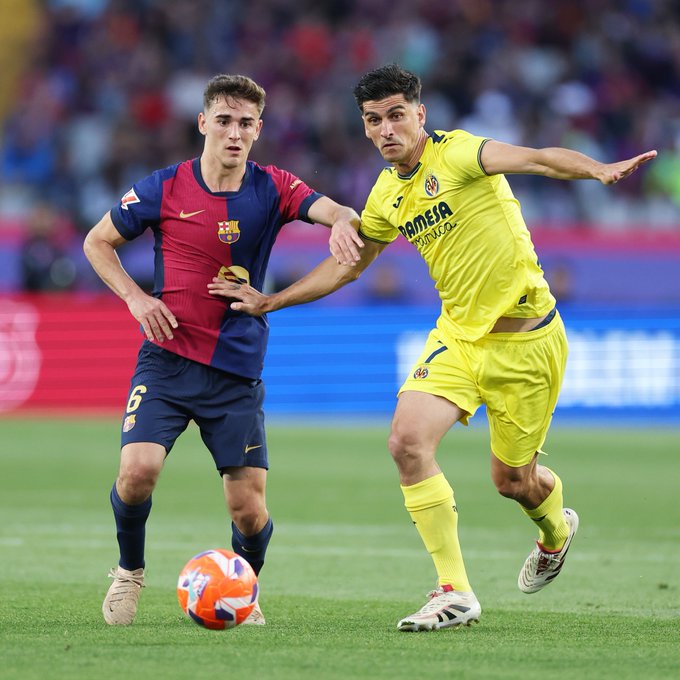 Dos jugadores de fútbol en acción en un campo verde durante un partido, uno con una camiseta roja y azul del Barcelona con el número 6 y el otro con un uniforme amarillo del Villarreal, ambos alcanzando una pelota naranja y blanca cerca de sus pies, con asientos de estadio y carteles publicitarios en el fondo.