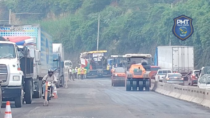 Wide view of a road construction site on a highway with multiple large trucks parked along the roadside, including a white semi-truck with cargo, yellow bulldozer, orange road roller compacting asphalt, and workers in orange vests and helmets standing near the equipment. A motorcycle with rider in helmet is positioned nearby, and traffic cones mark the work area. Lush green vegetation and hills line the background, with a blue shield sign displaying PMT logo visible on the right side.