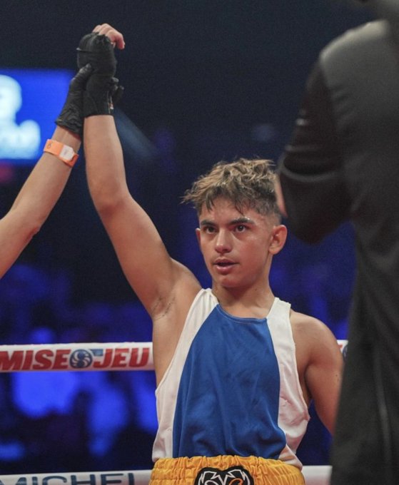 Un joven boxeador con el pelo despeinado se para en un ring de boxeo, levantando su brazo derecho con un guante negro en alto en aparente victoria, con la boca ligeramente abierta, vistiendo una camiseta sin mangas blanca con detalles azules, calzoncillos azules y blancos con el logotipo de un patrocinador y una pulsera naranja. Detrás de él, se ve una multitud borrosa y un árbitro con atuendo negro, con cuerdas iluminadas en azul y una pancarta de patrocinador en las cuerdas que dice Michelob Ultra. El escenario es una arena cubierta con iluminación tenue enfocada en el ring.