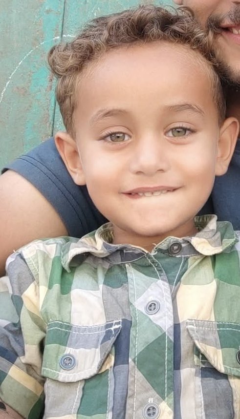 A young boy with curly brown hair and light skin smiles at the camera, showing his teeth, while held by an adult male with a beard visible in the frame. The boy wears a green and white checkered short-sleeved shirt. The background is a weathered green door or wall.