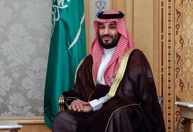 First image shows a man wearing traditional Saudi attire including red and white checkered headscarf and robe seated on ornate chair with hands clasped, Saudi green flag with sword emblem in background, wooden paneled room setting. Second image depicts an older man in dark suit white shirt blue tie gesturing with hands while speaking into microphone at podium, blue and white Israeli flag behind him, outdoor or stage setting.