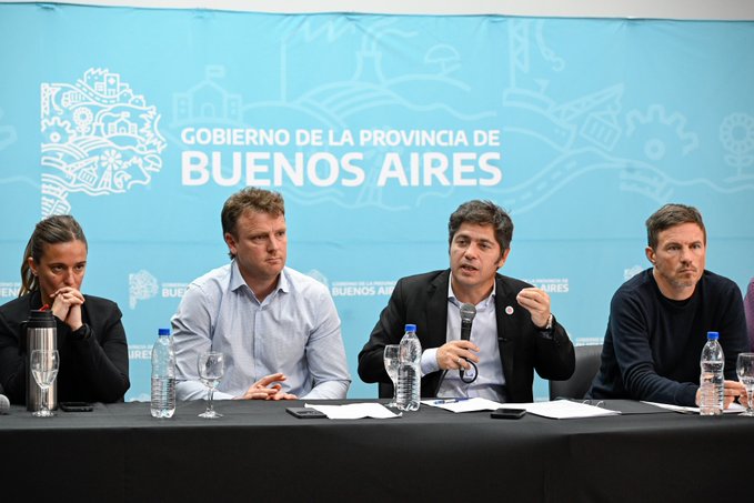 Four people sit at a long black table in front of a blue backdrop with white text reading Gobierno de la Provincia de Buenos Aires. A woman in a dark top holds a tumbler, a man in a light blue shirt sits with hands on table, another man in a dark suit gestures while speaking, and a man in a dark sweater sits with arms crossed. Bottles of water and glasses are on the table along with papers.