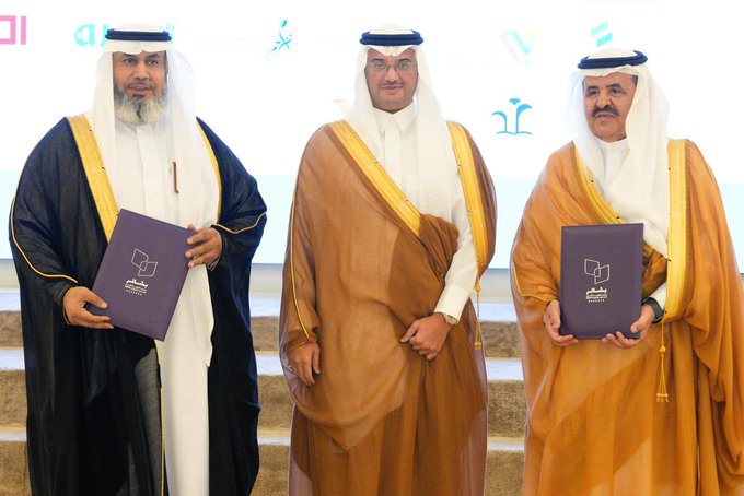 Three men in traditional Saudi thobes and headscarves stand side by side on a stage with a beige marble-like backdrop. The man on the left wears a dark thobe with gold trim and holds a purple folder with Arabic text. The middle man wears a light brown thobe and holds a similar purple folder. The right man wears a white thobe with gold trim and holds another purple folder. Blue and white logo-like elements appear on the backdrop.