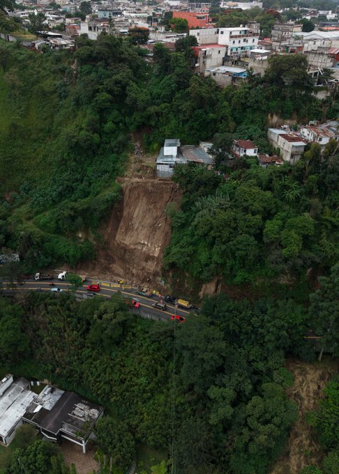 Aerial view of a hillside landslide blocking a road with vehicles stopped nearby surrounded by green vegetation and urban buildings in the background. Close-up of workers in yellow safety vests and helmets operating machinery to clear debris and install barriers along the sloped road edge. Group of workers including those wearing CIV-labeled shirts handling wooden beams and tools near the unstable soil slope with safety cones and barriers in place. Roadside scene with dump truck unloading materials while workers in helmets and vests set up wooden supports and barriers amid scattered debris on the asphalt.