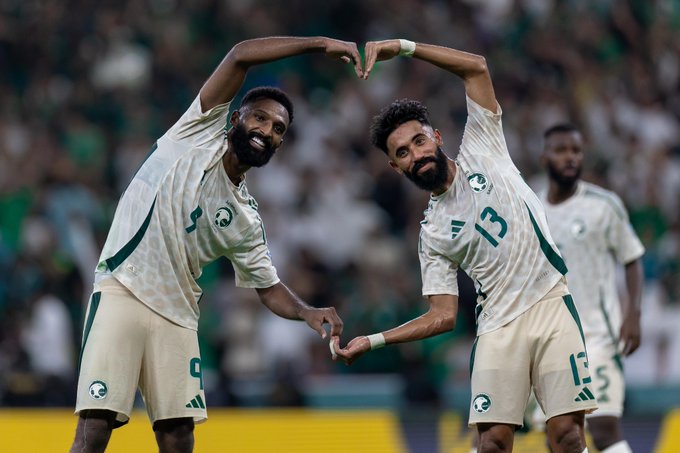 First image shows Faysal Fajr in a green and white Adidas Saudi Arabia national team jersey with number 9, placing his index finger over his mouth in a silencing gesture, standing on a football field in front of a white goal net with green posts, blurred stadium crowd in green attire visible in the background. Second image depicts Faysal Fajr in number 9 jersey and Saud Abdulhamid in number 13 jersey, both smiling and raising their arms to form a heart shape with hands touching at the bottom, wearing matching green and white patterned shorts and jerseys, on a field with blurred green-shirted spectators and yellow stadium barrier behind them.