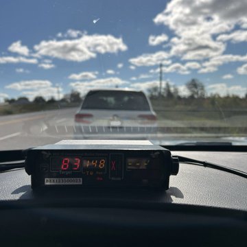 First image shows a dashboard-mounted speed radar device displaying 148 km/h with an X indicator inside a vehicle windshield with a white car visible ahead on a road under a partly cloudy blue sky. Second image depicts a white SUV on a flatbed tow truck on the roadside with a black police SUV parked nearby surrounded by autumn foliage trees and a clear road under sunny skies with scattered clouds.