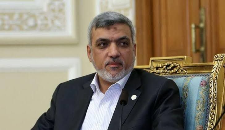 A middle-aged man with gray hair and a beard wearing a dark suit white shirt and a small pin on his lapel sits in an ornate wooden chair with gold accents and patterned upholstery in a room with wooden doors and decorative walls.