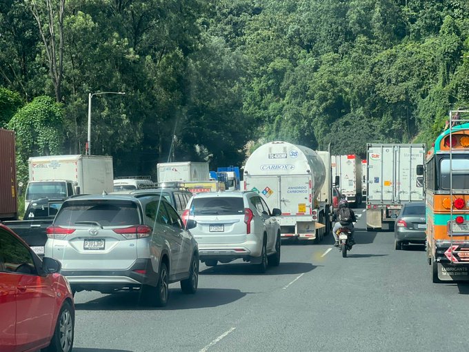 Multiple views of a busy highway scene with heavy traffic congestion including several large trucks stopped or slowed one behind the other a blue tractor-trailer with visible mechanical issues at the center a yellow tanker truck nearby various cars SUVs and buses in line motorcycles weaving through and lush green trees lining the roadside under clear daylight conditions