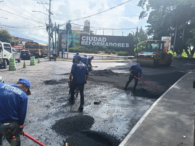 Several workers in blue uniforms and yellow vests are repairing a potholed asphalt road on a busy urban street, using shovels to spread fresh blacktop material while a yellow steamroller compacts it nearby. Traffic cones and barriers are placed around the work area for safety, with vehicles like a white truck and school bus visible in the background. A large green banner reading CIUDADADORES stretches across a fence, and palm trees line the sunny roadside setting.