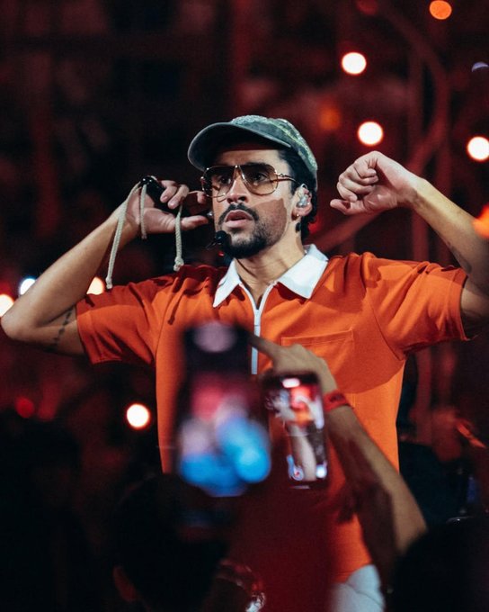 Bad Bunny performs on a brightly lit stage with orange and red lighting and hanging bulbs. He wears a green cap, dark sunglasses, an orange short-sleeve shirt with white collar, and holds a microphone. His arms are raised dynamically, with a wristband visible, and a cord hangs from the mic. Audience members in the foreground hold up phones recording the show.