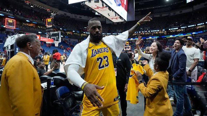 LeBron James in yellow Lakers jersey number 23 stands on basketball court with arms raised gesturing excitedly surrounded by fans in yellow attire and others in various clothing including suits and casual wear in arena with scoreboard and banners visible.
