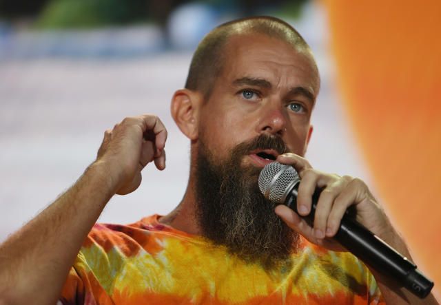 A bald man with a beard wearing an orange tie-dye shirt holds a microphone while gesturing with his hand on stage against an orange background.