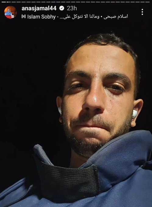 Close-up portrait of a young man with short dark hair, beard, and serious expression, wearing white wireless earbuds and a dark blue hoodie with a high collar, set against a plain black background. Arabic text overlay appears at the top in white, reading انجمال44 23ح, and additional text below including انس جمال في اسلام صبيح. Instagram watermark is visible in the bottom right corner with username anasjamal44.