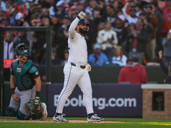 El jugador de béisbol con uniforme blanco con detalles azules levanta el brazo derecho en celebración en el campo, con guantes en la mano y casco de bateo, mientras que el receptor con ropa verde y azul se encuentra cerca sosteniendo el bate, la multitud con atuendo naranja visible en el fondo detrás de la red, el anuncio en la pared detrás.