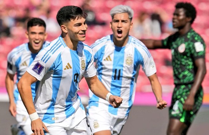 Un grupo de jugadores de fútbol masculinos con uniformes de la selección argentina con rayas azules y blancas y camisetas con los números 9 y 11 celebran un gol en un campo rojo del estadio, un jugador con cabello plateado con una camiseta verde de Nigeria cerca, asientos del estadio al fondo.