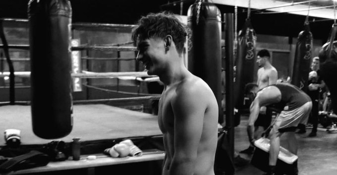 Fotografía en blanco y negro dentro de un gimnasio de boxeo con poca luz que muestra varios sacos de boxeo colgando del techo, un ring de boxeo en el fondo con cuerdas, un joven sin camisa con cabello rizado parado cerca del ring sonriendo mientras mira hacia otro lado, otro hombre sin camisa agachado cerca en pantalones cortos, figuras adicionales entrenando o de pie en el fondo, incluido uno levantando pesas, Equipo de gimnasio como botellas de agua y toallas en el piso.