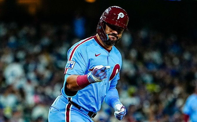 Kyle Schwarber se para en posición de bateo en un campo de béisbol durante un juego nocturno, con un casco rojo de los Filis con el logotipo de la P, una camiseta alternativa azul con rayas rojas, el número 12, muñequeras rojas y sosteniendo un bate. El uniforme presenta los logotipos de Nike y Fox Sports. Una multitud llena las gradas en el fondo bajo las luces del estadio.