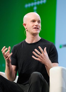 A bald man with fair skin wearing large hoop earrings and a black t-shirt sits on a white chair gesturing with both hands while speaking on stage against a green background with a white logo resembling a cross. A simple orange background features a large white Bitcoin symbol in the center.