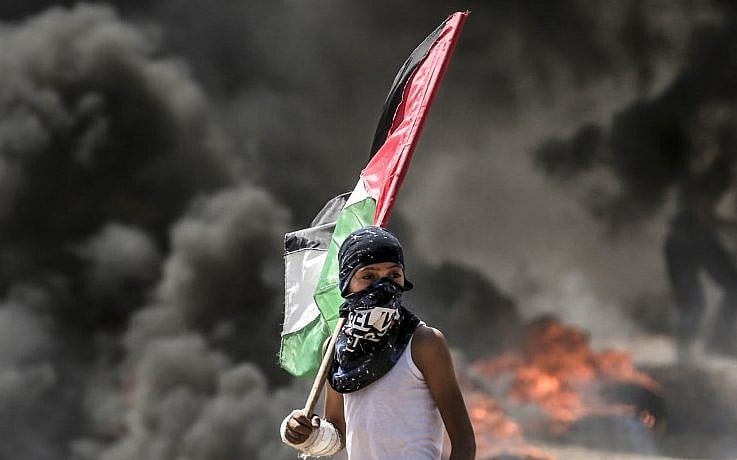 A young child with a black cloth covering their face stands holding a wooden staff topped with a Palestinian flag, dressed in a white tank top, positioned in front of thick black smoke billowing from fires on the ground in an outdoor urban protest environment.