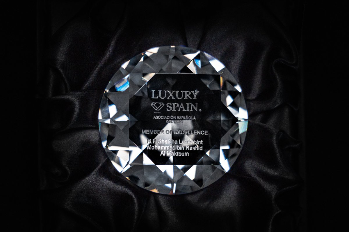 A woman wearing a black hijab and abaya sits at a desk in an office setting with a large digital screen displaying a colorful map in the background, looking towards the camera with a neutral expression. A crystal diamond-shaped award trophy engraved with Luxury Spain logo, Association of Excellence, Diamond Awards for Excellence, and Al Maktoum name rests on a black velvet cloth, featuring multifaceted surfaces and metallic accents.