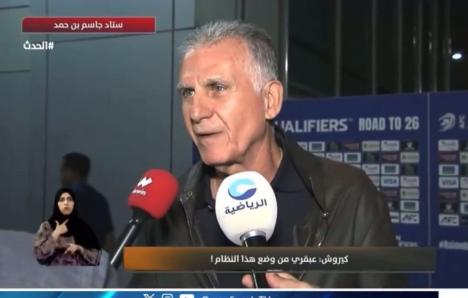 An older man with gray hair and glasses wearing a dark jacket speaks into a red microphone labeled Al Jazeera during a press conference. He stands in front of a backdrop with Asian Football Confederation logos and text reading QUALIFIERS ROAD TO 26. Another microphone with a Qatar logo is visible nearby. A woman in a black hijab and a man in a suit stand behind him holding microphones. The setting appears to be an indoor event space with bright lighting.