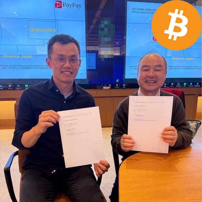 Two Asian men in business attire sit at a wooden table in a modern office holding identical white documents with text and signatures visible. The man on the left wears glasses and a black shirt smiling at the camera. The man on the right wears a gray sweater smiling while holding the papers. Behind them large screens display PayPay logos multiple Bitcoin symbols and text mentioning Bull Capital and SoftBank Capital. The setting includes wooden paneling and orange accents with Bitcoin icons.