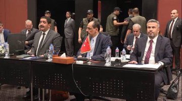 First image shows group of men in suits and traditional attire seated at long conference table with Turkish flag water bottles tissues and laptops in modern room with red walls and people standing behind. Second image depicts four bald or gray-haired men in suits sitting at conference table with water bottles tissues and documents in light-colored room with chairs. Third image features multiple men some in suits some in white thobes seated around L-shaped conference table with documents water bottles flowers and abstract painting on wall in carpeted room with plants. Fourth image captures group of men including some in thobes and suits shaking hands near chairs with water bottles flowers and abstract painting in background.