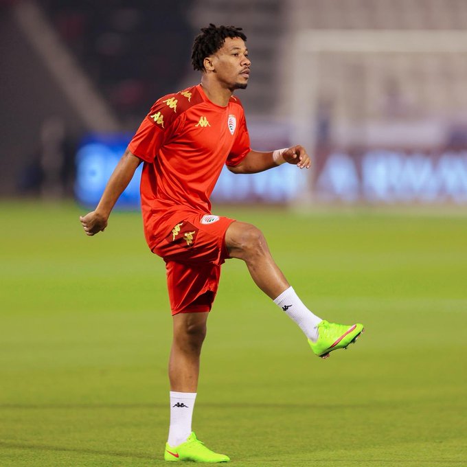 Player Jameel Al-Yahmadi stands on green soccer field at night stadium with dark background and stadium lights. He wears red Kappa uniform jersey and shorts with sponsor logos, white Nike socks, and green Nike cleats. His pose shows one leg lifted as if in motion during play, with focused expression.