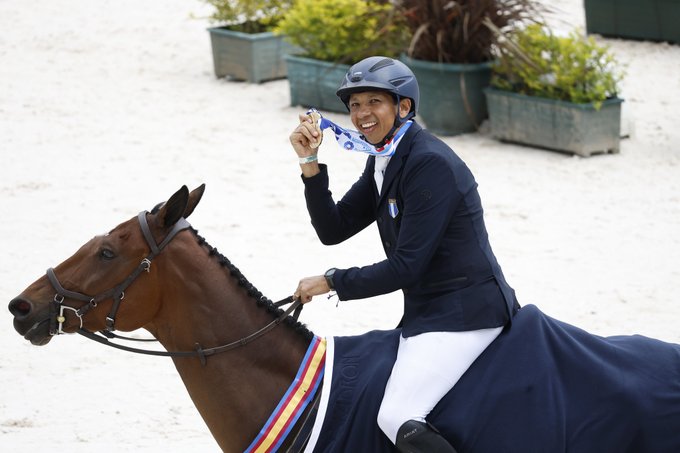 A male equestrian rider in a navy blue jacket, white pants, and helmet sits on a brown horse covered with a multicolored sash, holding a blue ribbon medal in his right hand while smiling. The horse stands on a sandy arena with green plants in pots in the background. Another rider in similar attire is visible nearby on a different horse.