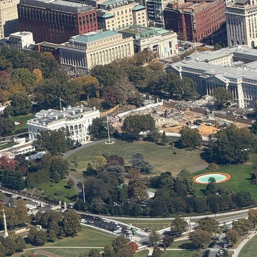 Aerial view of the White House surrounded by trees with autumn leaves green lawns a circular fountain and parked cars the East Wing shows construction with debris scaffolding and partial demolition of the building facade nearby structures include domed capitol-like buildings and urban architecture