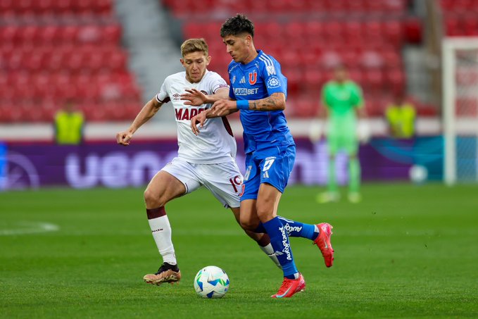 Two soccer players in action on a green field during a match one in white and maroon kit number 19 from Lanús team dribbling the ball while another in blue kit number 10 from Universidad de Chile defends closely nearby a goalkeeper in green uniform stands in the goal area red stadium seats and advertising boards visible in the background including sponsor logos like Adidas and others the scene captures intense competition near the penalty area with the ball in play.