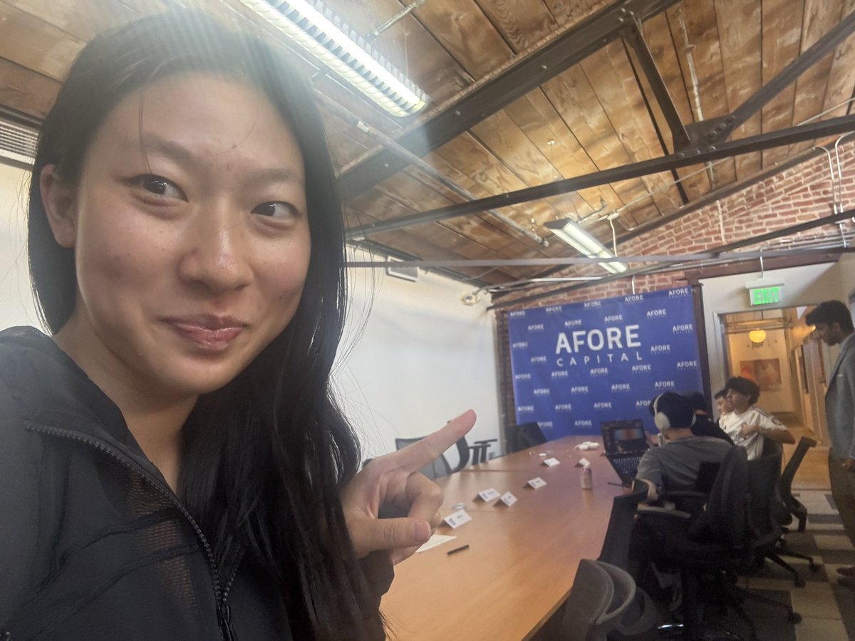 A selfie-style photo of an Asian woman with long dark hair, wearing a black zip-up hoodie, pointing directly at the camera with her right index finger while smiling slightly; she is seated or positioned near a wooden conference table in a modern industrial office with exposed brick walls, wooden beam ceiling, fluorescent lighting, and several people (including men in casual attire with headphones) sitting around the table using laptops and papers; a large blue banner with 'AFORE CAPITAL' logo dominates the background, along with name tags and coffee cups on the table.