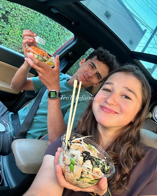 Two young adults sit in the front seats of a parked car with a green hedge visible outside the open sunroof. The man on the left wears a light blue shirt and holds a bowl of salad with a fork, smiling slightly. The woman on the right wears a gray tank top and holds a bowl of poke with chopsticks, smiling broadly. Both bowls contain rice, vegetables, tofu, and other ingredients. A watch is on the mans wrist and seatbelts are visible.