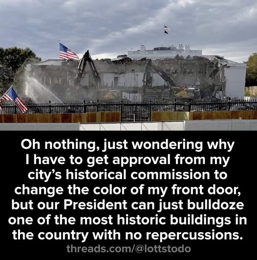 The White House building partially demolished with gray debris piles and exposed structure visible. Two yellow excavators positioned in front with one arm extended and another spraying water on dust. American flags on poles in foreground and background. Black metal fence with barriers and orange construction netting enclosing the site. Overcast sky above trees.