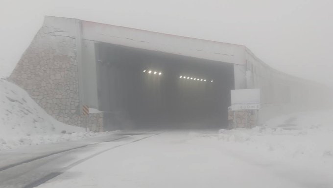 Snow-covered road leading to a large tunnel entrance with bright lights inside visible through fog and heavy snow accumulation on brick walls and ground surrounding the structure under overcast sky