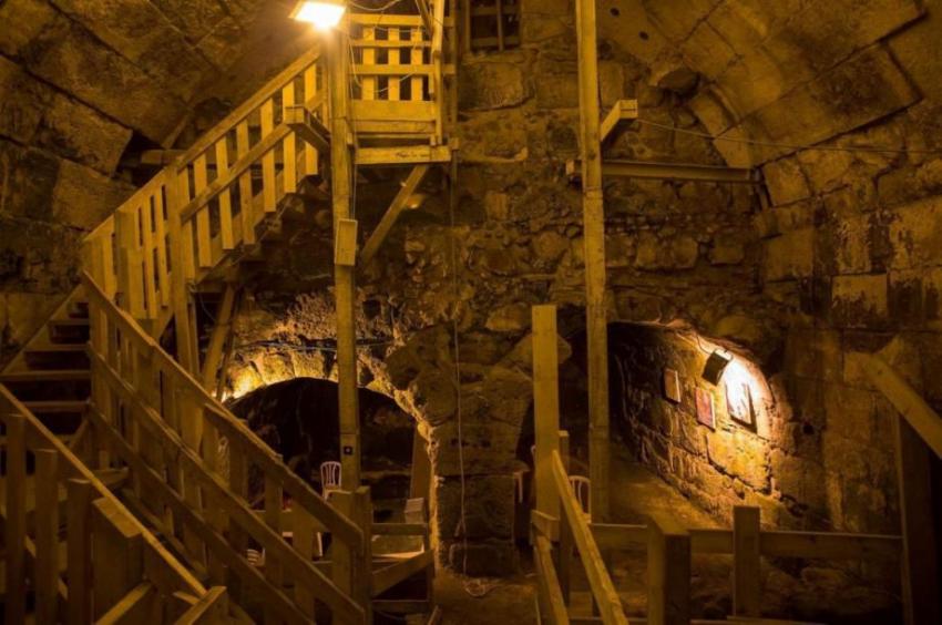 Underground stone cavern with arched walls and ceilings, featuring wooden railings and stairs leading down into dimly lit tunnels, illuminated by hanging lamps casting yellow light on the rocky surfaces and structural supports.