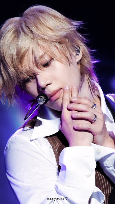 Blonde-haired man with earpiece and rings on fingers holds microphone close to mouth, wearing white shirt and brown vest, gazing sideways with neutral expression under purple stage lights.